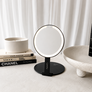 A black LED vanity mirror with a circular illuminated frame and a slim stand, styled on a light beige surface with books, a candle, and a bowl in a bright interior setting.