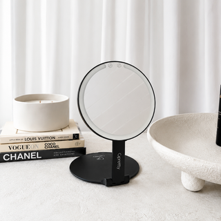 A black round vanity mirror with a slim stand and built-in LED lighting, styled on a light textured surface next to a ceramic candle, books, and a bowl against a white curtain background.