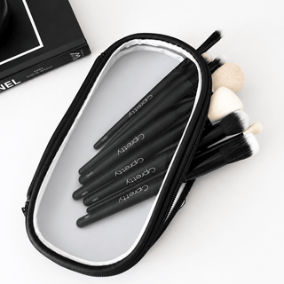 A black zippered cosmetic bag with a clear front panel, containing several black makeup brushes with white and beige bristles. The bag is photographed on a white surface next to a black book.